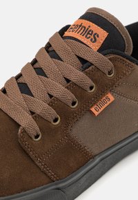 Sapatilhas de camurça castanha e lona com pormenores em preto, atacadores texturizados e uma etiqueta laranja com o logótipo "etnies". Sola de borracha e ilhós metálicos visíveis.