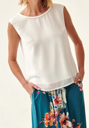 Ärmellose weiße Bluse mit geflochtenem Ausschnitt, kombiniert mit türkisfarbenen Hosen mit großen bunten Blumenmustern, Hände in den Taschen.