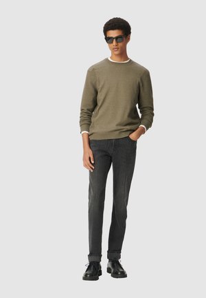 Olive green textured sweater with white trim, paired with dark jeans and black lace-up shoes. The model stands with hands in pockets, wearing sunglasses.
