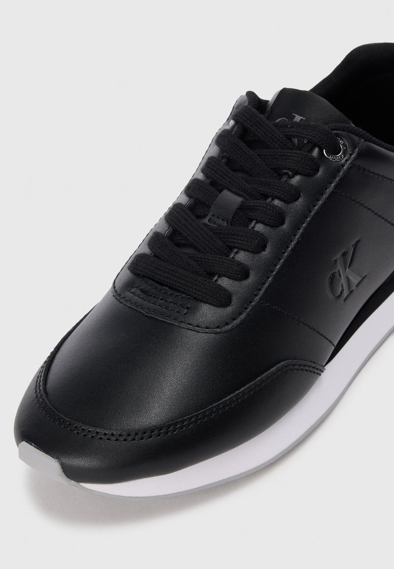 Zwarte leren sneaker met zwarte veters en witte zool, voorzien van reliëf "CK" logo aan de zijkant.