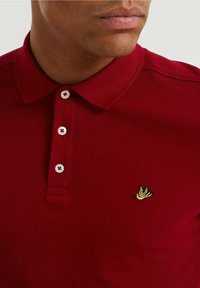 Röd polo t-shirt i texturerat tyg, med en klassisk krage, tre vita knappar och en liten gul fågel-logotyp p å bröstet.