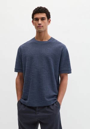 Mann mit lockigem Haar, der einen strukturierten marineblauen Kurzarmpullover und dunkle Hosen trägt, steht mit den Händen in den Taschen.