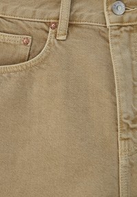 Lysbrun kordfløjlsstof med en fint ribbet struktur. Har en lomme, kobbernitter og en sølvknap med branding.