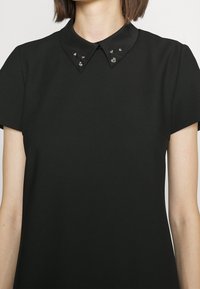 Femme portant une robe noire à manches courtes avec un col pointu orné de petites épingles décoratives en argent.