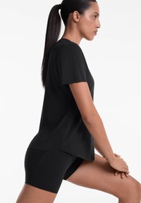 Black short-sleeve top with a loose fit, made from smooth fabric; paired with black fitted shorts. Side pose showing posture and design.