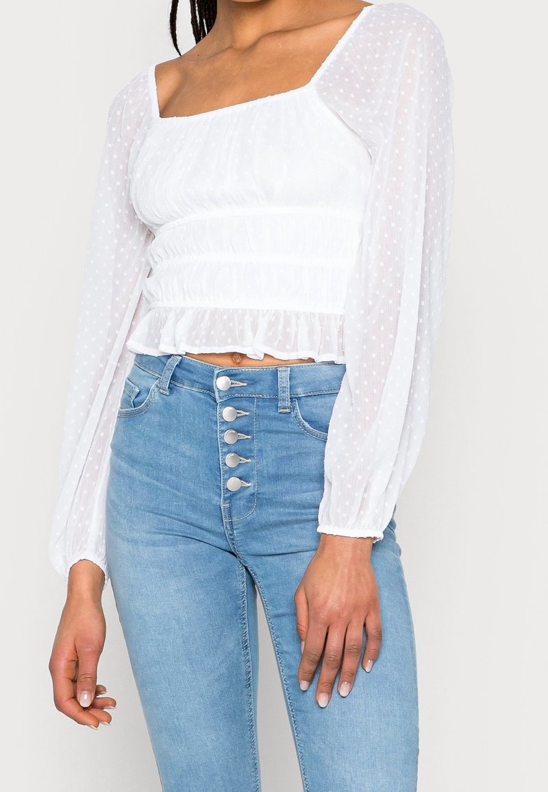 Kvinna i en vit genomskinlig blus med långa ärmar och prickar, tillsammans med ljusblå högmidjade jeans med fem silverknappar.