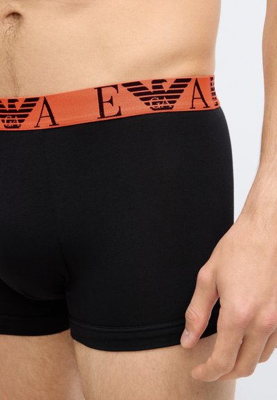 Boxer in cotone nero con una fascia elastica arancione che presenta un logo nero. Tessuto liscio, design aderente e lunghezza fino a metà coscia.