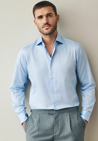 Chemise bleu clair à manches longues avec un col classique, associée à un pantalon gris sur mesure avec des plis à l'avant et des poches latérales.