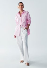 Camicia rosa con colletto e maniche lunghe, abbinata a pantaloni bianchi larghi e infradito beige. Tessuto semplice senza motivi distintivi o accentuazioni.