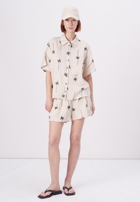 Lyse beige skjorte og shorts sett med svart palmetrasjé. Kortermet skjorte med knapping foran; settet inkluderer en matchende caps. Svarte flip-flops.