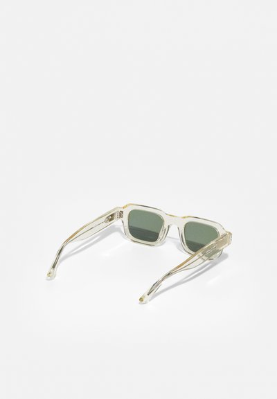 Gafas de sol transparentes con montura cuadrada, lentes verdes, brazos transparentes delgados y sutiles acentos dorados en los laterales.