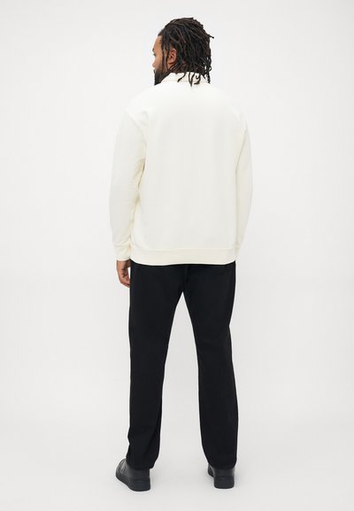 Άνδρας με κοτσιδάκια που φοράει λευκή πουλόβερ, μαύρα παντελόνια και μαύρα παπούτσια, στέκεται με την πλάτη απέναντι σε απλό φωτεινό φόντο.