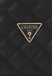 Sac à main matelassé noir avec un écusson en logo triangulaire doré portant "GUESS" et un point d'interrogation, fabriqué en matériau synthétique.