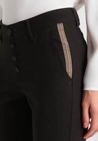 Svarta byxor har en texturerad tyg, en rad svarta knappar på midjan och en sidoficka med en metallisk mönstrad accent.