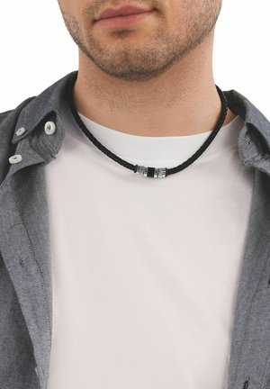 Primo piano di un uomo che indossa una collana intrecciata nera con dettagli in argento sopra una camicia bianca e una camicia grigia sbottonata.