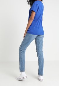 Blå bomullströja med korta ärmar, kombinerad med ljusblå slitna jeans och vita högskodda sneakers, sedd bakifrån.