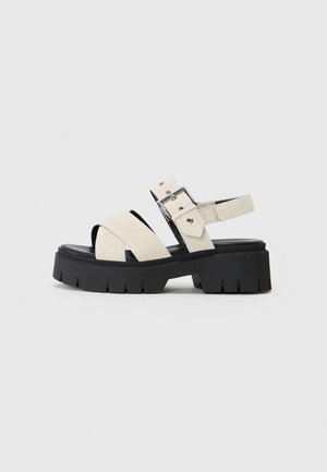 Platform sandals featuring a textured black rubber sole, cream suede straps, and a silver buckle accent on a cross-strap design.