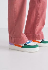 Multicolored sneakers with a green and white upper, white rubber sole, and accents of purple, orange, and blue; paired with pink pants.