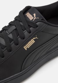 Black leather sneaker with a textured finish, featuring black laces, a gold Puma logo on the side, and a low-profile rubber sole.