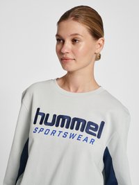 Ung kvinde med lys hud og samlet hår iført en hvid sweatshirt med navyblåt "hummel SPORTSWEAR"-logo, der kigger let mod højre.