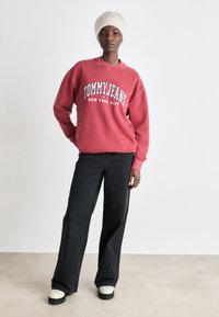 Crvena dukserica s otisnutim "TOMMY JEANS NEW YORK CITY", kombinirana s crnim hlačama širokih nogavica i krem čizmama na platformu. Nošena je teksturirana krem beanie kapa.