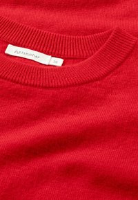 Maglione rosso lavorato a maglia con scollatura rotonda a coste. L'etichetta mostra "hessnatur" e taglia 36. Tessuto liscio con un colore uniforme.