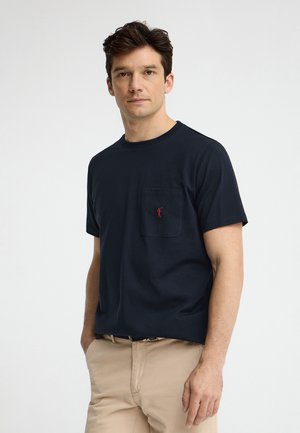 Mand med mørkt hår iført en navyblå kortærmet skjorte med et rødt logo på brystlommen og beige bukser, stående med hænderne i lommerne.