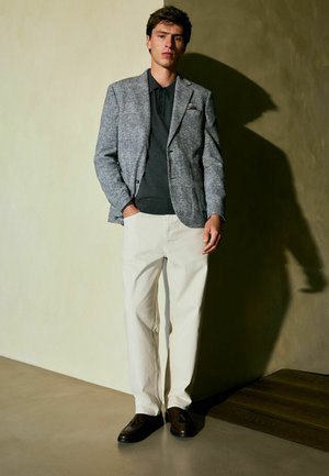 Blazer texturat gri peste un polo negru, asortat cu pantaloni off-white și pantofi maro închis. Accesoriu notabil - batista de buzunar. Fundal uniform.