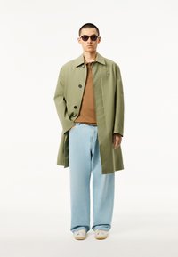 Olivgrön trenchcoat över en brun polo t-shirt, kombinerad med ljusblå vida jeans och vita sneakers. Mörka solglasögon fulländar looken.