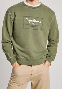 Hombre con sudadera oliva de Pepe Jeans London y pantalones beige, con las manos en los bolsillos, de frente, recortado por debajo de la nariz.