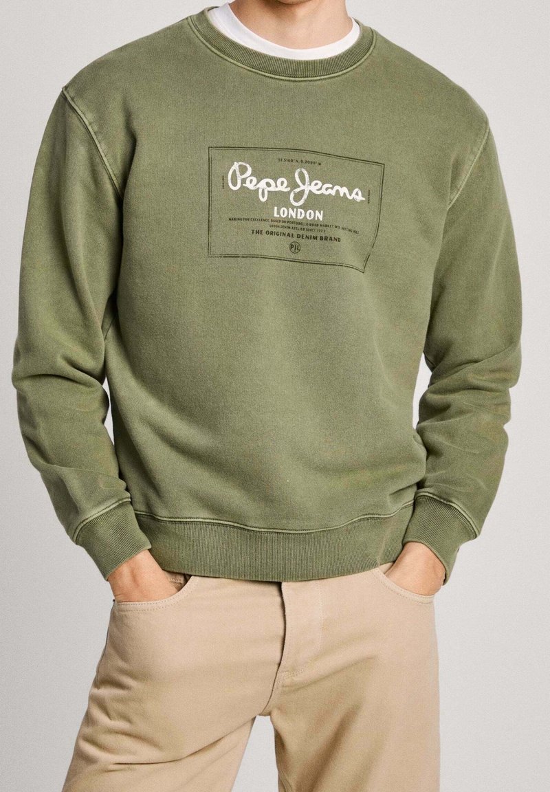 Hombre con sudadera oliva de Pepe Jeans London y pantalones beige, con las manos en los bolsillos, de frente, recortado por debajo de la nariz.