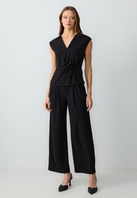 Zwart en gouden gestreepte twee-delige outfit met een mouwloze wikkeltop met een draai detail en wijde broekspijpen. Gladde stofstructuur.