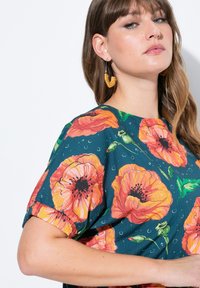 Blomsterkjole med store orange valmueprint på en mørk teal baggrund, korte ærmer og rund halsudskæringer; parret med gyldne hoops øreringe.