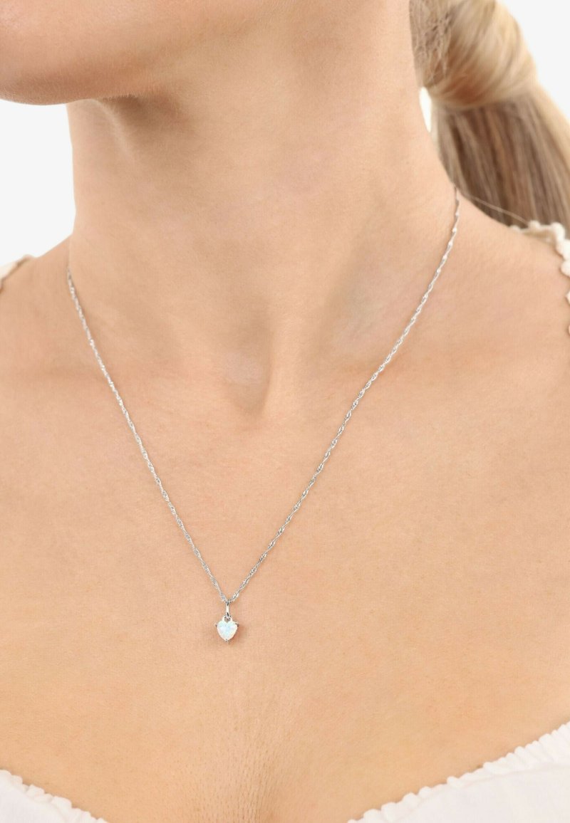 Collar de cadena de plata con un colgante en forma de corazón que presenta una pequeña gema, colocado sobre un tono de piel neutro. Diseño simple y delicado.