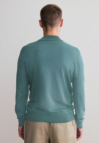 Myntegrøn pullover med lange ærmer og ribbet krave, der har en glat tekstur og tætsiddende pasform, parret med beige bukser.