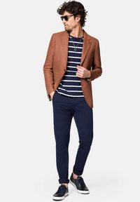 Brązowy, teksturowany blezer na granatowej, paskowanej koszuli, w połączeniu z granatowymi, dopasowanymi spodniami i czarnymi sneakersami. Dodatki obejmują okulary przeciwsłoneczne i naszyjnik.