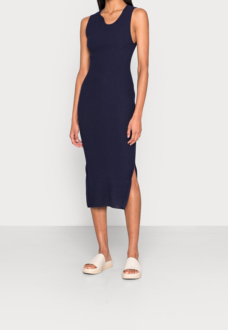 Robe midi bleu marine sans manches en maille côtelée avec fente latérale portée par une personne debout sur un sol gris clair devant un mur blanc.