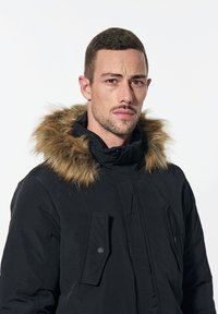 Parka nera con cappuccio foderato in pelliccia, dotata di due tasche anteriori e una texture liscia. Il design è isolato e funzionale.