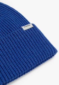 Gorro de punto acanalado azul hecho de material suave, con una pequeña etiqueta blanca con la palabra "SCALPERS" cosida en el borde doblado.