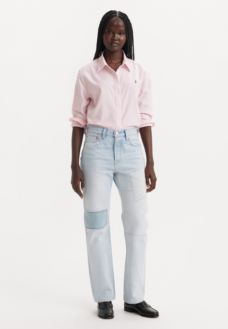 Camicia button-up rosa chiaro con maniche arrotolate. Jeans di denim azzurro chiaro con dettagli a toppe. Scarpa nera completa l'outfit.