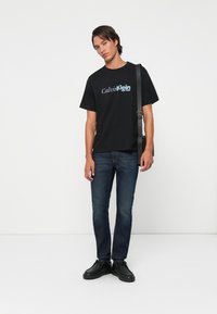 Mladý muž stojí, má na sobě černé tričko s logem Calvin Klein, tmavě modré džíny, černé boty a černou tašku přes rameno.
