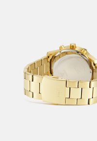 Reloj de metal dorado con pulsera de acero inoxidable, que presenta un diseño elegante, textura suave y un broche grabado con "GUESS."