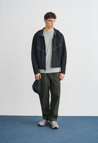 Czarna kurtka na zamek z szarymi akcentami, szara koszulka, oliwkowozielone spodnie i szare sneakersy; trzyma czapkę z daszkiem w kolorze czarnym. Prosty, funkcjonalny design.