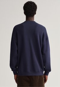 Námořnická modrá mikina vyrobená z měkkého materiálu, s kulatým výstřihem a volným střihem. Zadní strana má hladkou texturu a minimalistický design.
