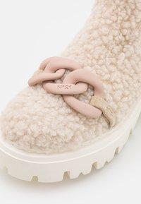 Gros plan sur une botte beige texturée avec une semelle blanche épaisse et un grand détail de chaîne rose portant l'inscription "N°21" à l'avant.