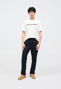 Camiseta blanca con el logo negro "JACK & JONES", combinada con pantalones deportivos azul marino oscuro y zapatillas marrones. Diseño sencillo y casual, ajuste recto en las piernas.