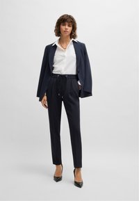 Marineblauwe op maat gemaakte blazer over een witte blouse met kraag, gecombineerd met zwarte broek met trekkoord en zwarte puntige hakken. Gladde stofstructuur.
