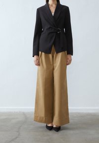 Mustat bleiserit, joissa on kääntödesign ja hopeinen rengasviimeistely, yhdistettynä korkeavyötäröisiin, leveälahkeisiin beigenvärisiin housuihin. Sileä kankaan rakenne.