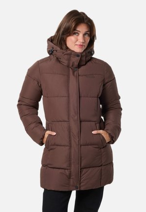 Une femme présente un manteau d’hiver marron, matelassé et à capuche, les mains dans les poches, sur un fond uni et clair.