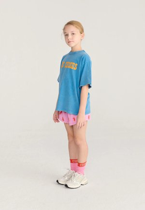 Jeune fille debout portant un T-shirt bleu oversize, un short rose avec des imprimés rouges, des chaussettes roses, des bandes rouges et des baskets blanches sur un fond uni.